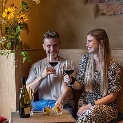 Ein Mann und eine Frau in schicker Kleidung stoßen mit Rotwein an und lächeln. Stimmungsvolle Atmosphäre in der Vinothek Knapp.