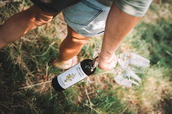 Die Beine einer Person, sie läuft über eine sommerliche Wiese und hält eine Flasche Wein mit Etikett in der Hand. Natur trifft Genuss und Rätsel.