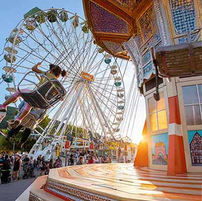 Besucher gehen durch einen belebten Rummelplatz mit Riesenrad und Fahrgeschäften während der Michaelismesse.