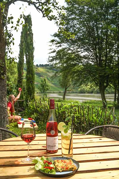 Startpunkt Outdoor Escape Kröv – Ein gedeckter Holztisch im Garten mit einer Flasche Rosé vom Weingut Hahn, Limonade und einem Teller mit Speisen. Im Hintergrund grüne Weinreben.