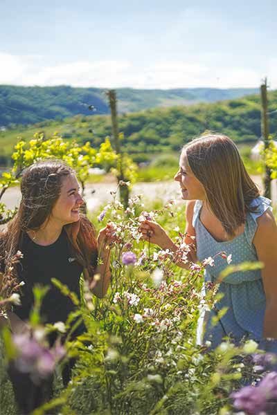 Zwei Frauen in einer Wildblumenwiese reichen sich strahlend zwei Blümchen – ein Moment voller Freude im Sonnenschein.
