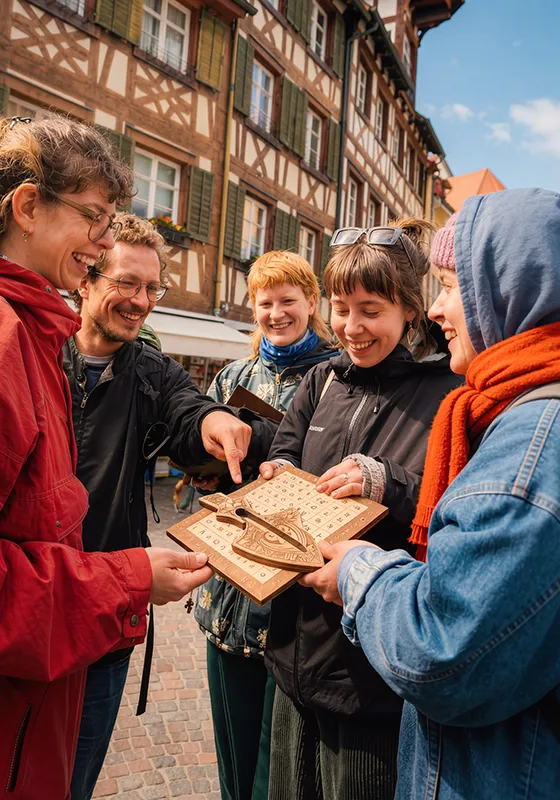 4 Freunde stehen in Meersburg zusammen und schauen auf ein Rätselgegenstand. Die Sonne strahlt ins Bild.